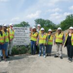 A group of people wearing white hard hats and safety vests stand pose in front of a construction site sign.