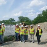 A group of people wearing white hard hats and safety vests stand pose in front of a construction site sign.