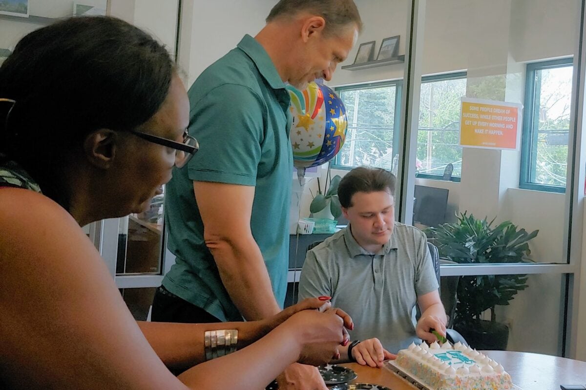 A man cuts a cake with white icing as another man and woman watch.