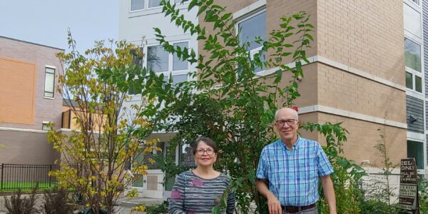 Man and woman pose in front of a tall green bush in front of an apartment building.