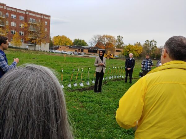 A woman wearing a headcovering speaks in a field with a line of shovels sticking in the dirt behind her.