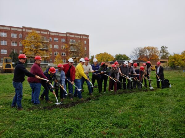 People wearing hard hats shovel dirt in a field. A large apartment building is in the background.