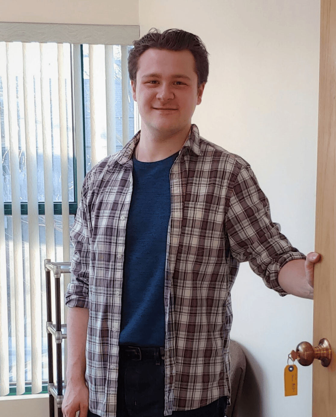 A white man with brown hair, wearing a navy shirt under a red, white and navy checkered shirt smiles as he holds open a door.