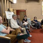 A group of people sitting in chairs in a circle listening to one man talking.