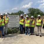 A group of people wearing white hard hats and safety vests stand pose in front of a construction site sign.