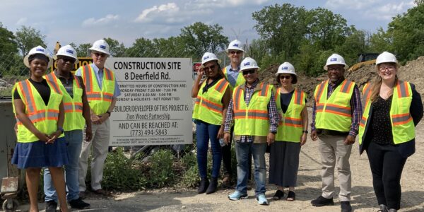 A group of people wearing white hard hats and safety vests stand pose in front of a construction site sign.