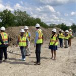 A group of people wearing white hard hats and safety vests stand on a construction site.