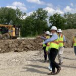 A man and woman gesture to others wearing white hard hats and safety vests stand on a construction site.