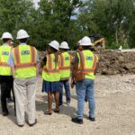 A group of people wearing white hard hats and safety vests stand on a construction site.