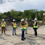 A group of people wearing white hard hats and safety vests stand on a construction site.
