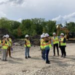 A group of people wearing white hard hats and safety vests stand on a construction site.