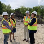 A group of people wearing white hard hats and safety vests stand talking on a construction site.