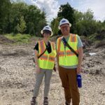 A white woman and a white man smile, wearing hard hats and bright construction safety vests.