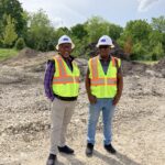 Two black men smile, wearing hard hats and bright construction safety vests.