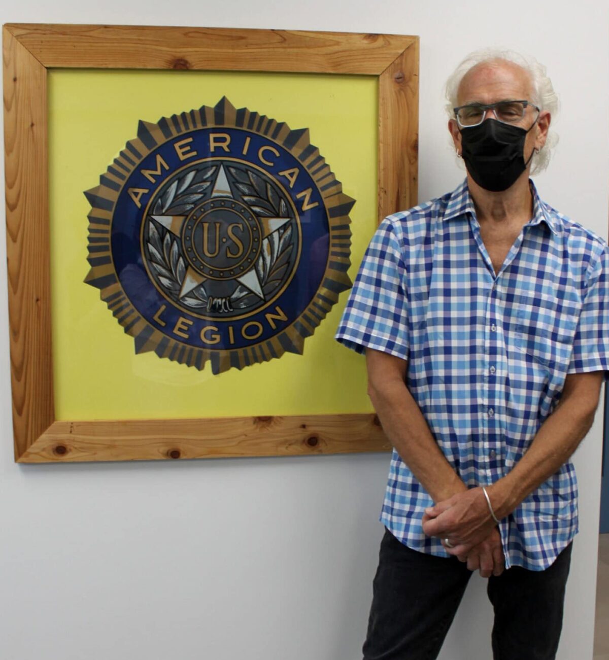 A man wearing a mask and a blue checkered shirt stands next to a wall hanging of a large American Legion logo.