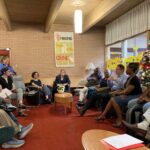 A group of people sitting in chairs in a circle listening to one man talking.