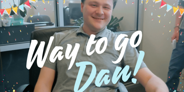 A white man with brown hair and waring a grey polo shirt poses in front of a cake with white icing. "Way to go Dan!"