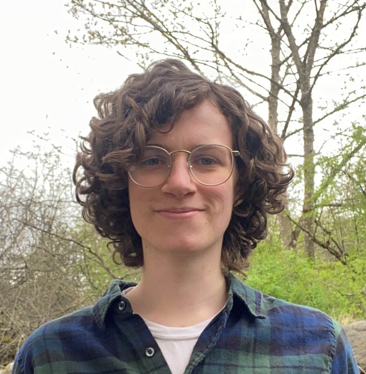 Young white woman with curly brown hair, wearing glasses, white t-shirt and blue/green plaid shirt.