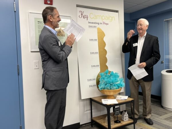 Two men smile and gesture to a poster with a large gold key on it.