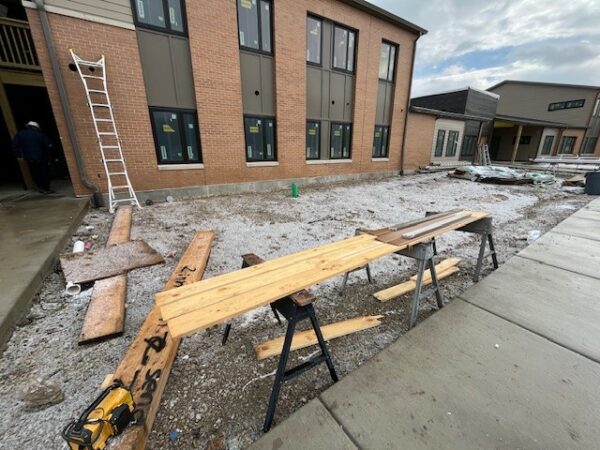 A construction site with wood laid across sawhorses ready to be cut. A two-story apartment building with brown brick in the background.