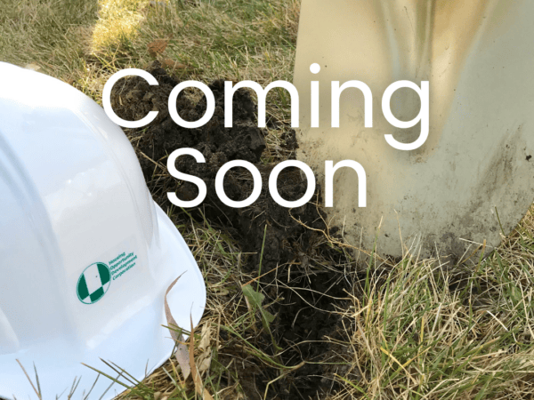 Hard hat and golden shovel on either side of a dirt hole surrounded by grass. White text that says "Coming Soon"