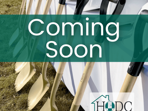 Golden shovels lean against a table covered with a white tablecloth with a row of white hard hats. The sun shines with grass in the background. "Coming Soon" in white text on a transparent green banner.