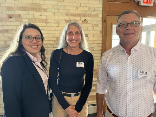 Two white women and one white man pose together. Light brick wall and wood accents in the background.