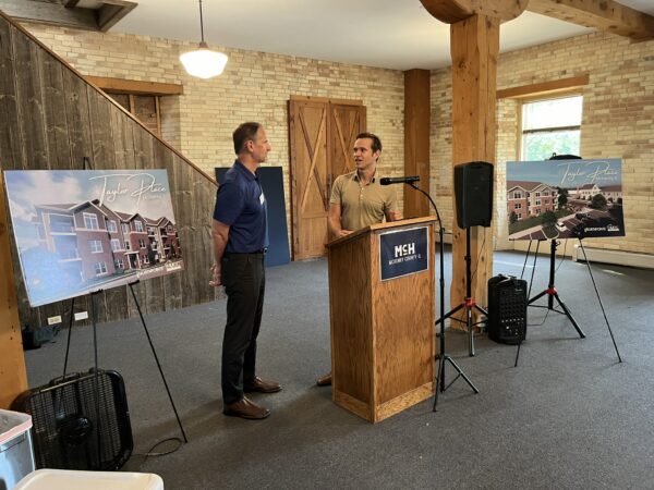 A white man wearing a tan polo shirt speaks from a podium as a white man wearing a navy polo shirt looks on.