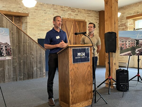 A white man wearing a navy polo shirt speaks from a podium as a white man wearing a tan polo shirt looks on.