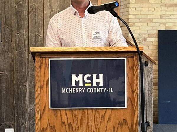 A white man wearing a light button-down shirt speaks from a podium.