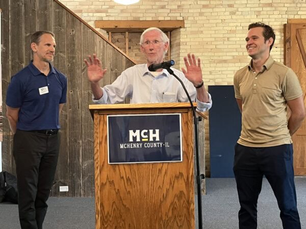 A white man wearing a light button-down shirt speaks from a podium as two white men look on.