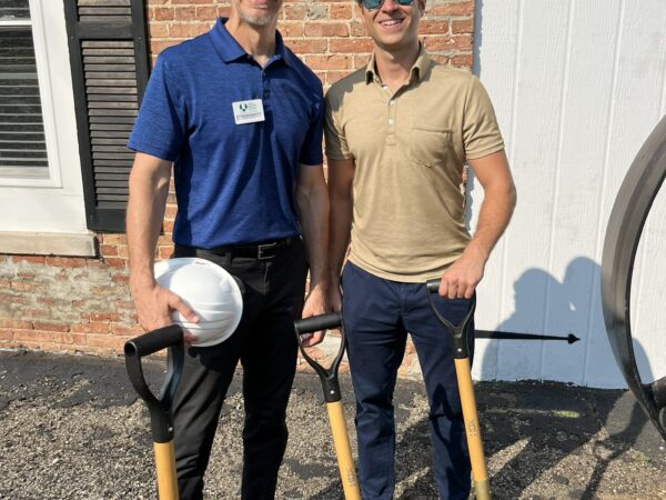Two white men wearing polo shirts and white hard hats pose with shovels in the dirt.
