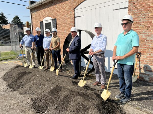 People wearing white hard hats pose with shovels in a mound of dirt in front of a brick building with a large iron wheel in the background.