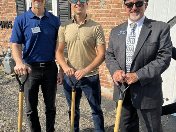 Three white men wearing white hard hats pose with gold shovels.
