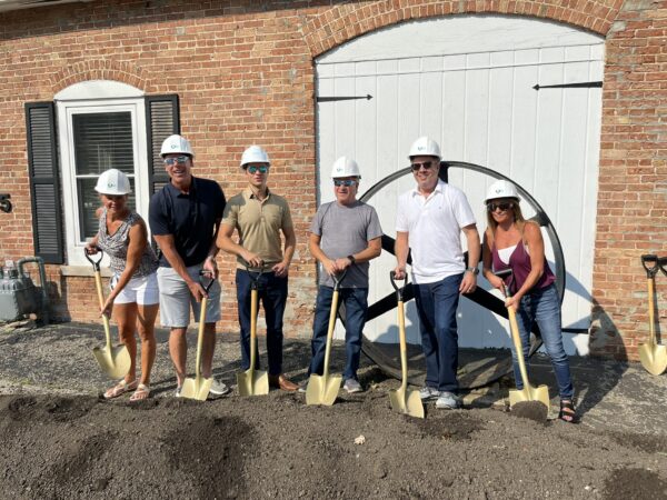 People wearing white hard hats pose with shovels in a mound of dirt in front of a brick building with a large iron wheel in the background.