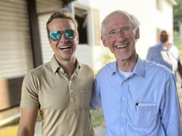 Two white men wearing summer shirts and one wearing sunglasses pose outside.