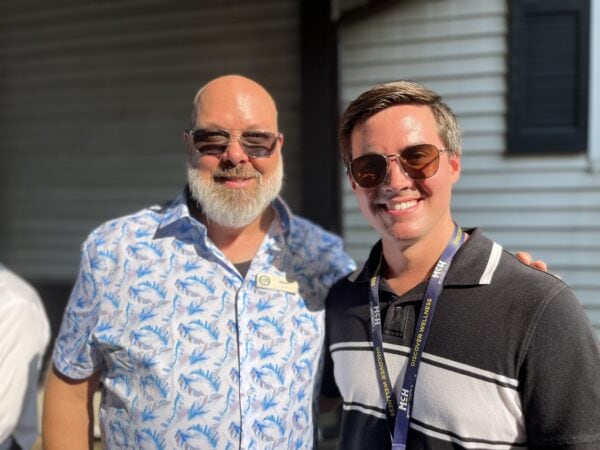 Two white men wearing summer shirts and sunglasses pose outside.