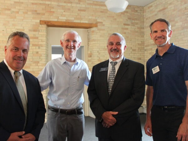 Group of white men wearing suits, or light shirts pose in a room with light brick walls and wooden accents.