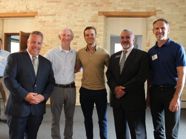 Group of white men wearing suits, or light shirts pose in a room with light brick walls and wooden accents.