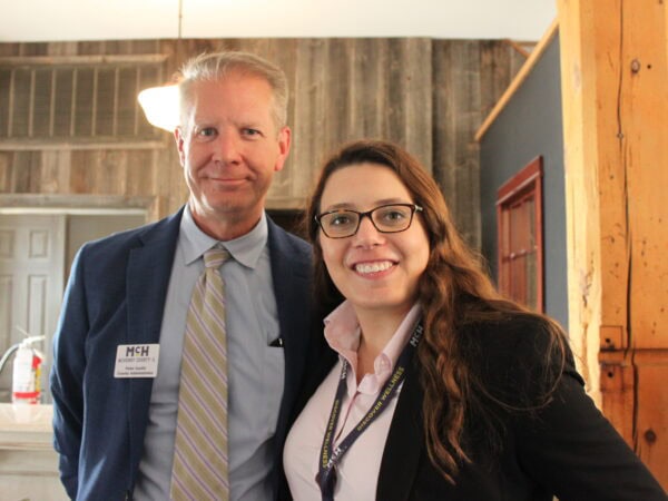 White man wearing a dark blazer with a light blue shirt and tie poses with a white woman with long brown hair, wearing glasses, a dark blazer and pink shirt.