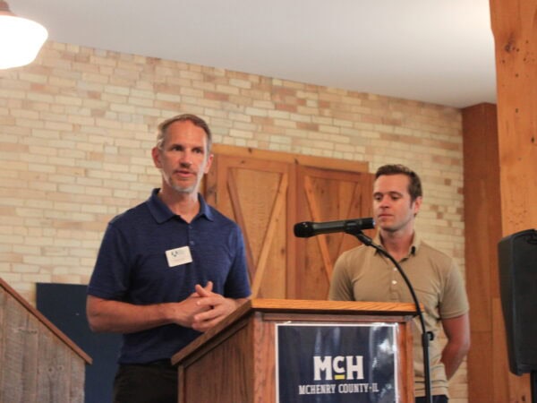 White man wearing a navy blue polo shirt talks in front of a podium while a man in a tan polo shirt looks on. A light brick background with oak wood doors.