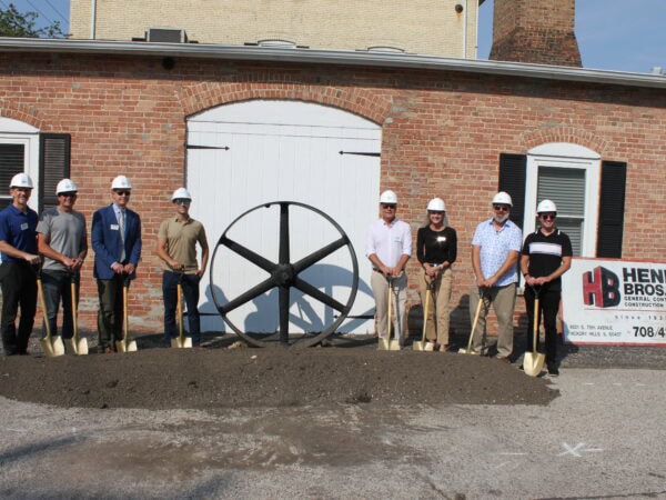 People wearing white hard hats pose with shovels in a mound of dirt in front of a brick building with a large iron wheel in the background.