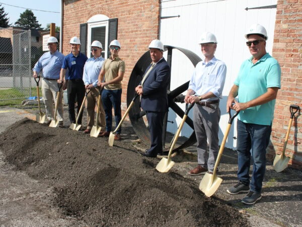 People wearing white hard hats pose with shovels in a mound of dirt in front of a brick building with a large iron wheel in the background.