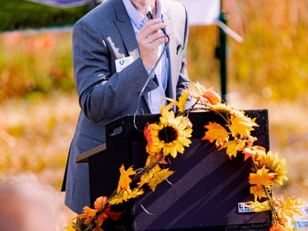 A white man wearing a blue blazer and light blue shirt speaks at a podium set up outside.