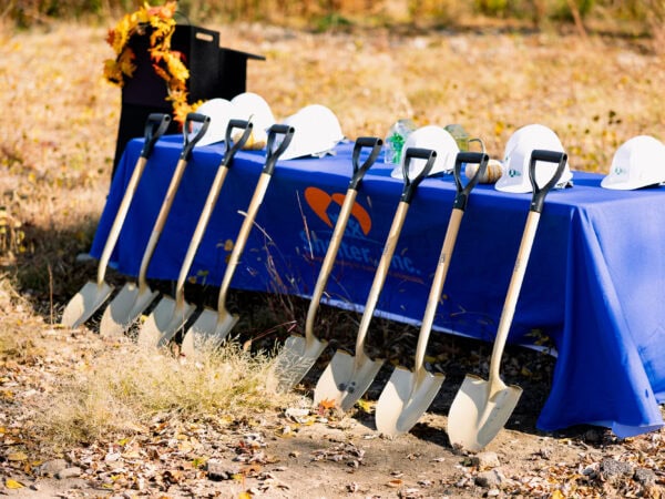 Shovels lean against a table with a blue tablecloth with white hard hats arranged. A podium is on the side of the table and adorned with yellow sunflowers.