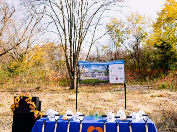 Shovels lean against a table with a blue tablecloth with white hard hats arranged. A podium is on the side of the table and adorned with yellow sunflowers. A construction banner is in the background surrounded by trees and grass.
