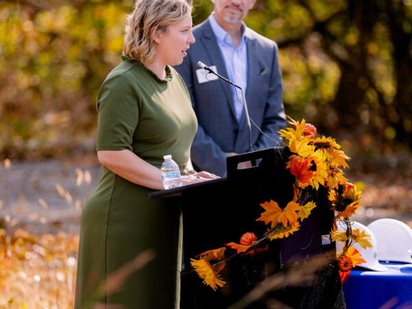 A white woman with blond hair and wearing a green dress speaks at a podium set up outside while a white man wearing a blue blazer and light blue shirt looks on in support.