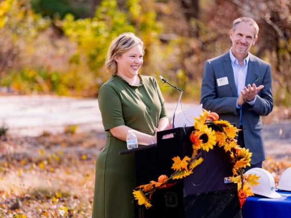 A white woman with blond hair and wearing a green dress speaks at a podium set up outside while a white man wearing a blue blazer and light blue shirt applauds.