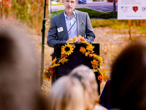 A white man wearing a blue blazer and light blue shirt speaks at a podium set up outside.