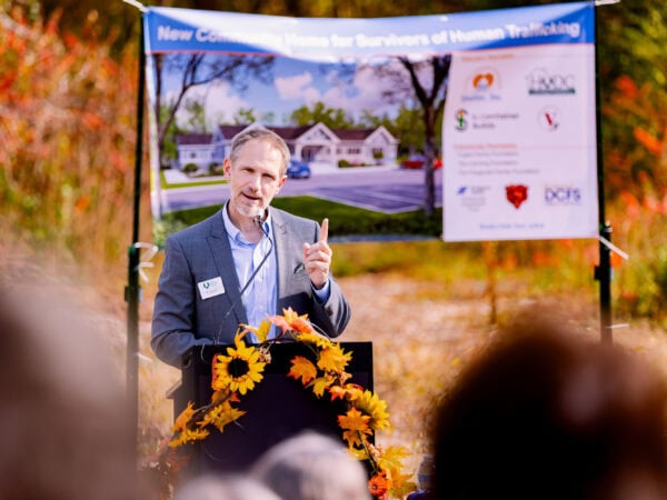 A white man wearing a blue blazer and light blue shirt gestures as he speaks at a podium set up outside.